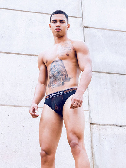 Man wearing a supawear neon mesh brief underwear against a light stone wall.