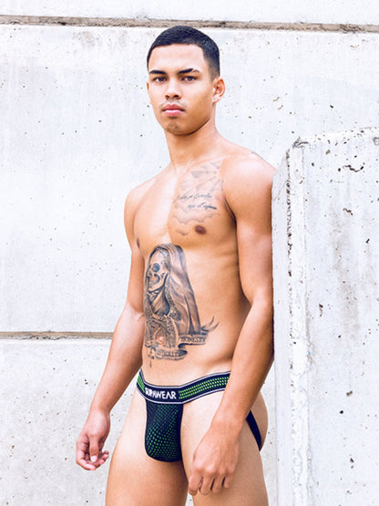 Man wearing a black jockstrap with green accents against a light gray wall.