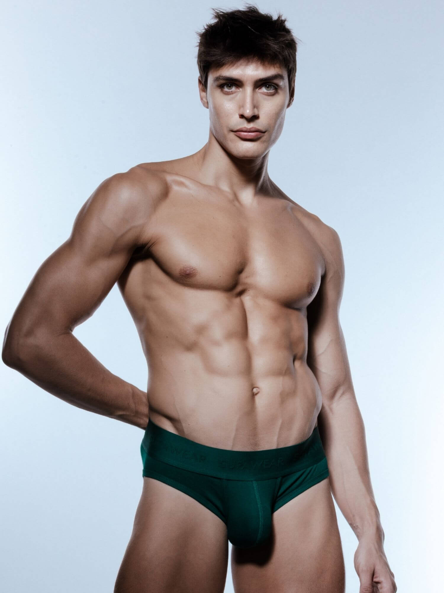 A muscular man wearing dark green briefs, posing against a light blue studio background.
