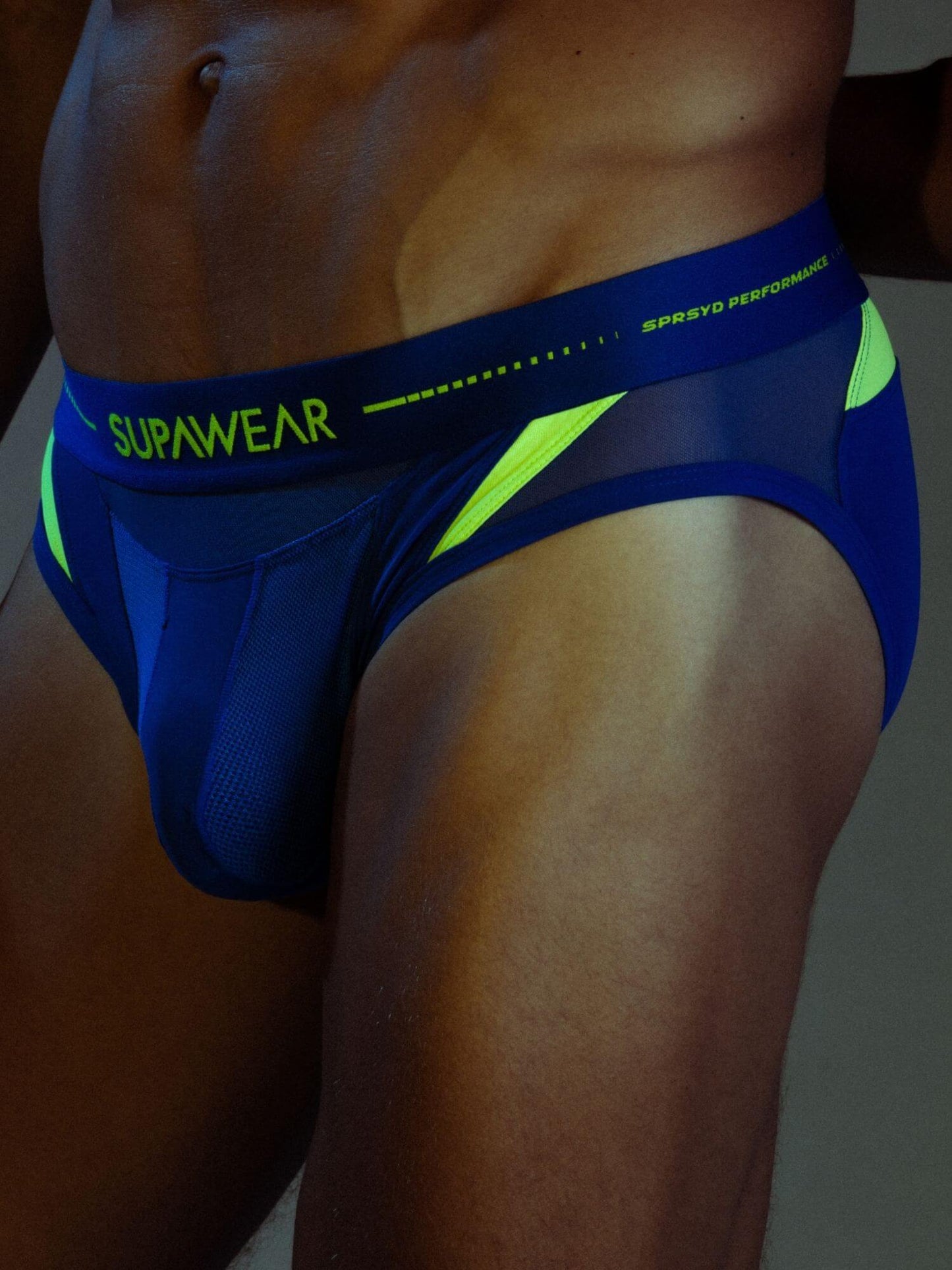 Close-up of a man wearing blue and neon yellow athletic briefs with the brand name SUPAWEAR visible on the waistband.