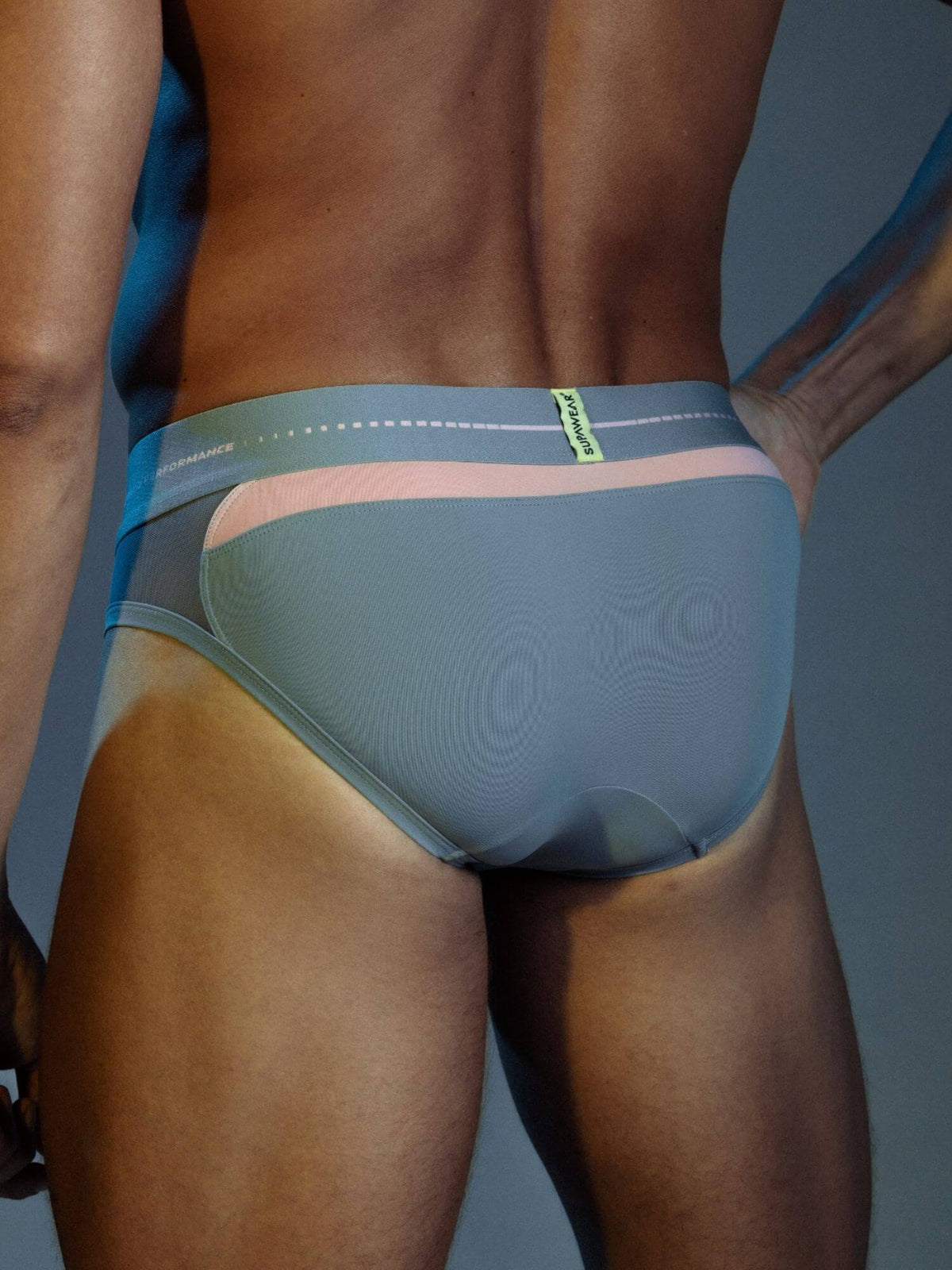 Rear view of a man wearing low-rise briefs with a textured slate blue main body, a light pink waistband accent, and a blue side panel with a small green brand tab.