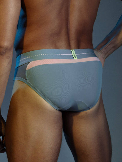Rear view of a man wearing low-rise briefs with a textured slate blue main body, a light pink waistband accent, and a blue side panel with a small green brand tab.