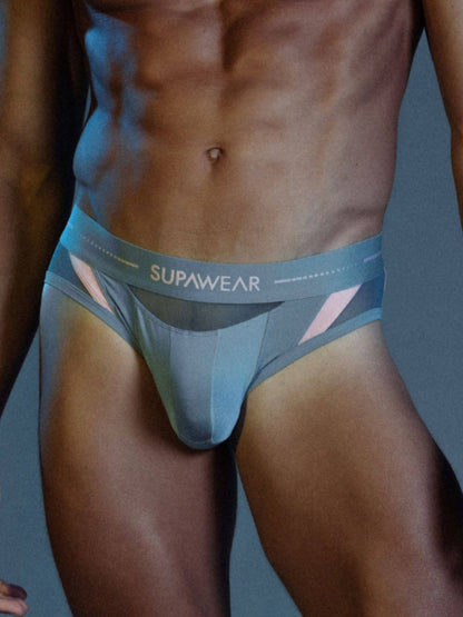 A close-up of a male model's lower torso wearing Supawear athletic briefs. The underwear is light blue-gray with a darker gray waistband featuring the 'SUPAWEAR' logo in white, and pink geometric side accents.