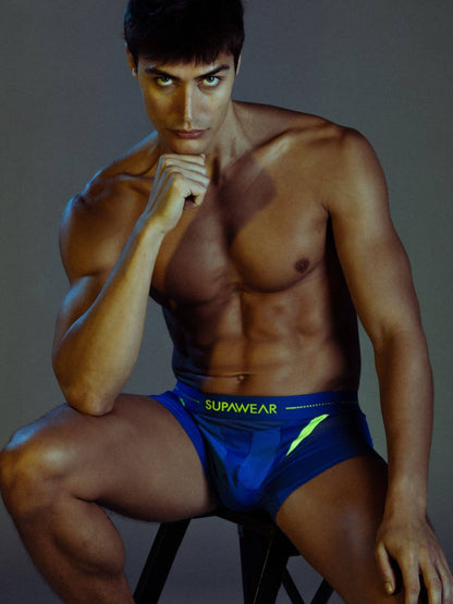 A muscular man wearing blue and green Supawear boxer briefs, posing on a stool against a dark gray studio background.