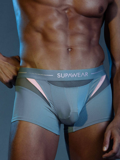 A close-up of a muscular male model wearing SUPAWEAR men's boxer briefs in a muted blue-gray color with pink and dark gray side accents.