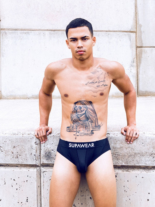 Man wearing black underwear with 'Supawear' branding against a stone wall.