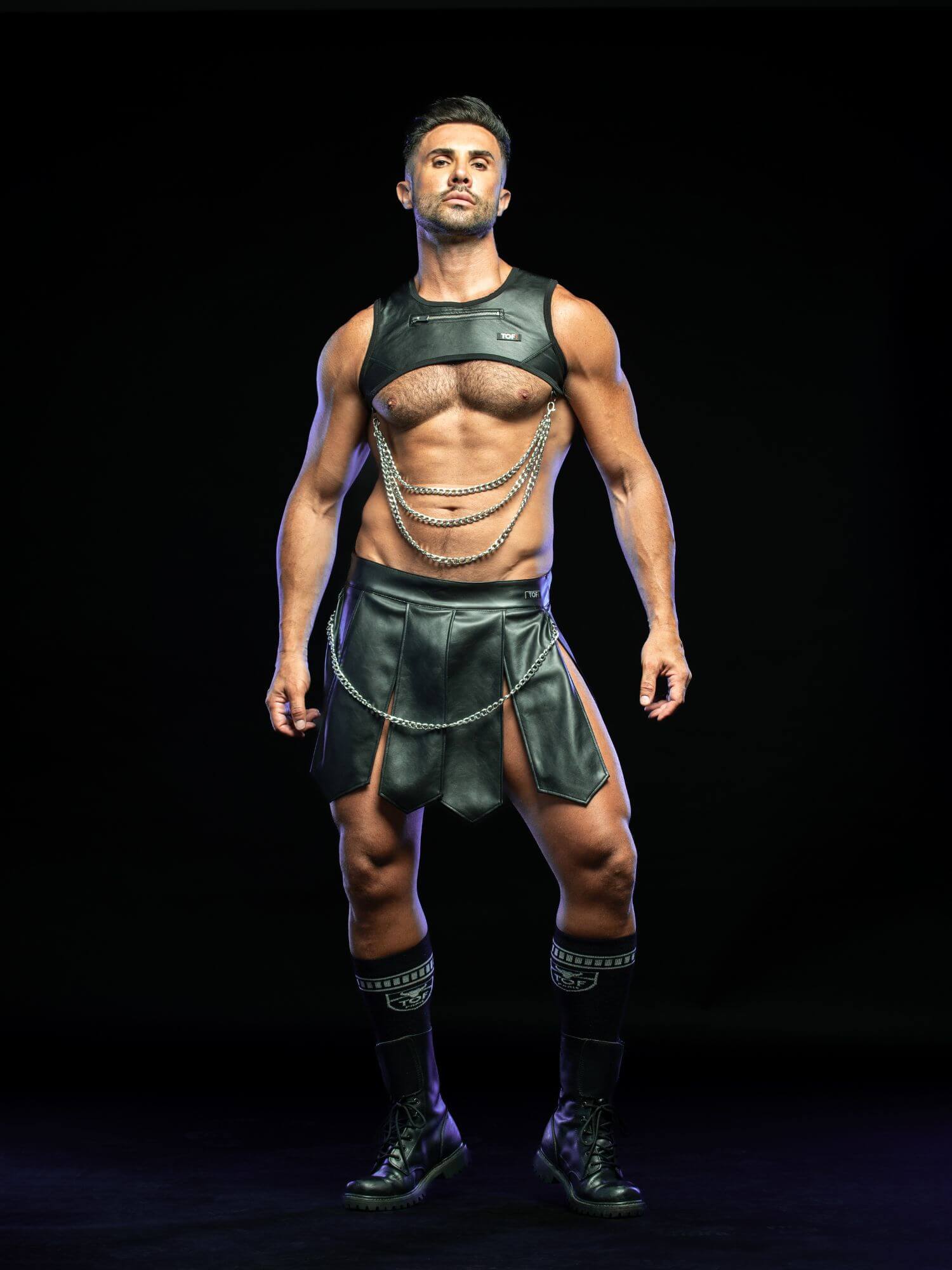 A muscular man modeling a black leather-style costume set consisting of a sleeveless top, matching skirt, and layered chain details, standing against a solid black background.