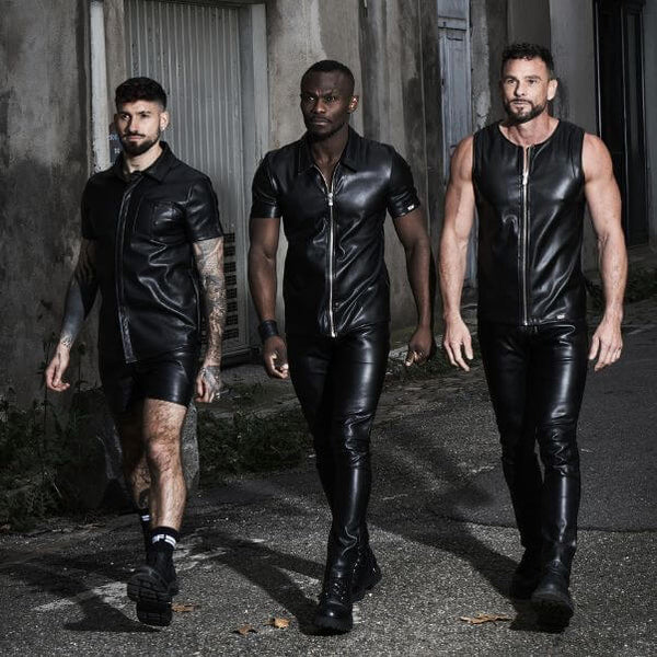 Three men in black leather outfits walking together against a textured wall.