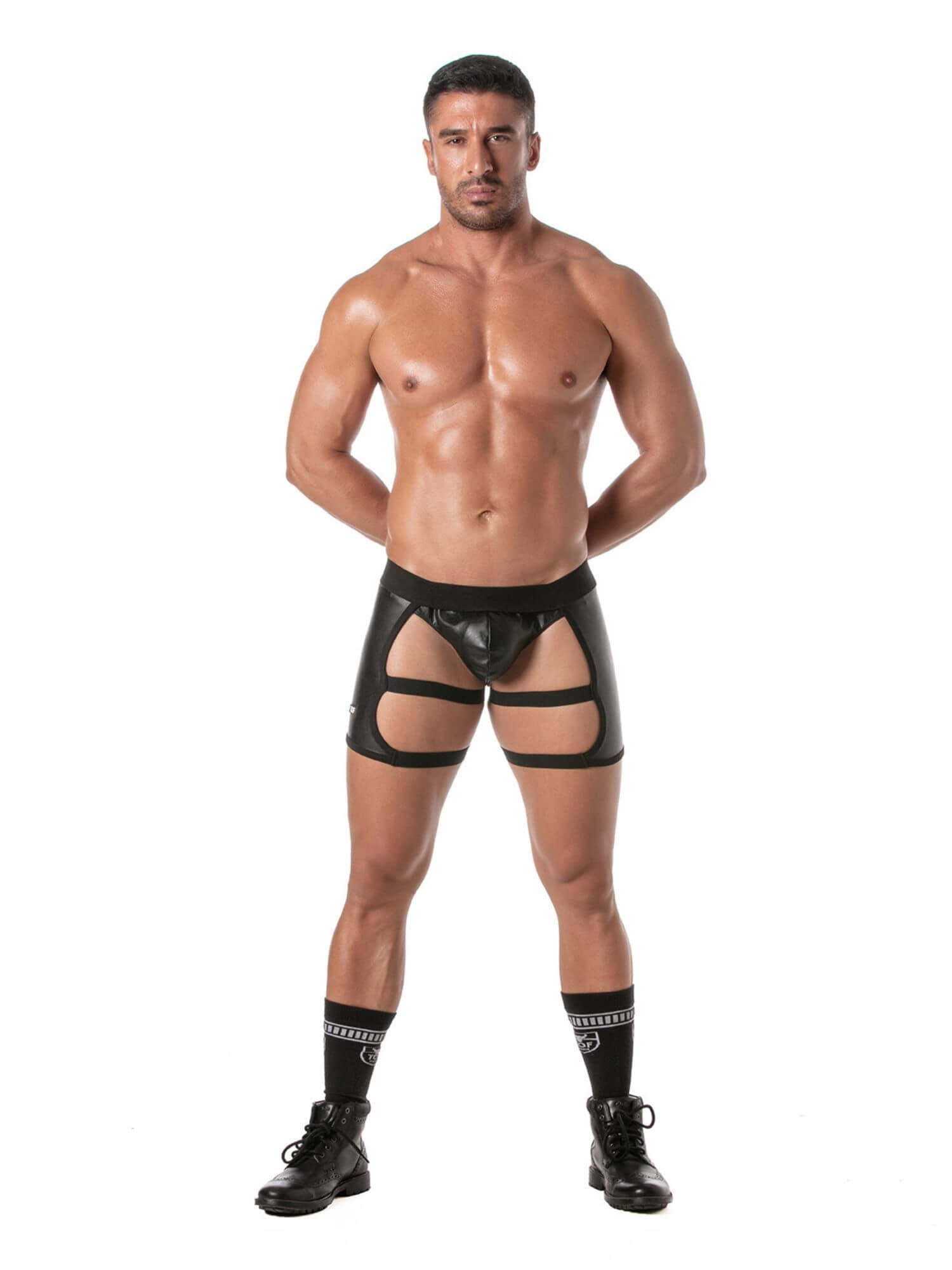 A muscular man modeling black glossy vinyl briefs with thigh garter straps and a front pouch, paired with black crew socks and black boots, against a solid white background.