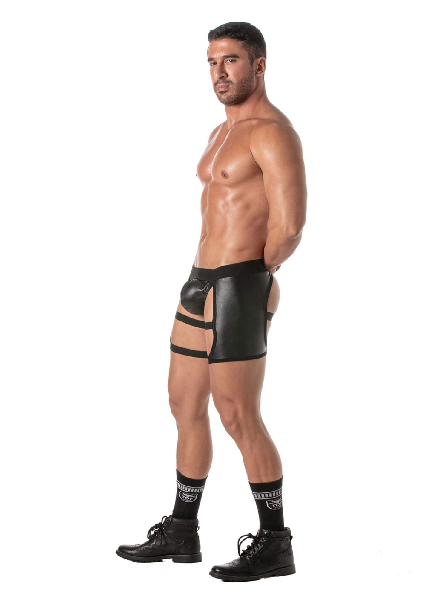 A man shown in profile wearing black strappy harness-style briefs, black socks, and black lace-up boots against a plain white studio background.