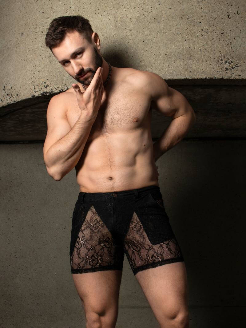 Man wearing black lace underwear against a textured wall.