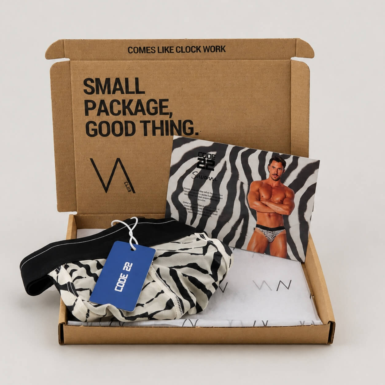 An example of a VA CLUB subscription box on a light grey background containing a CODE 22 Savanna print thong with black and white animal print, a blue CODE 22 brand tag, VOCLA branded tissue paper and a small printed product insert card