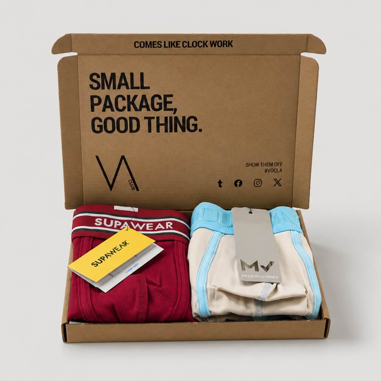 An example of a VA CLUB subscription box on a light grey background containing a burgundy SUPAWEAR Varsity boxer and a MODUS VIVENDI Dynamic beige and light blue brief with brand tags, VOCLA branded tissue paper and a small printed product insert card