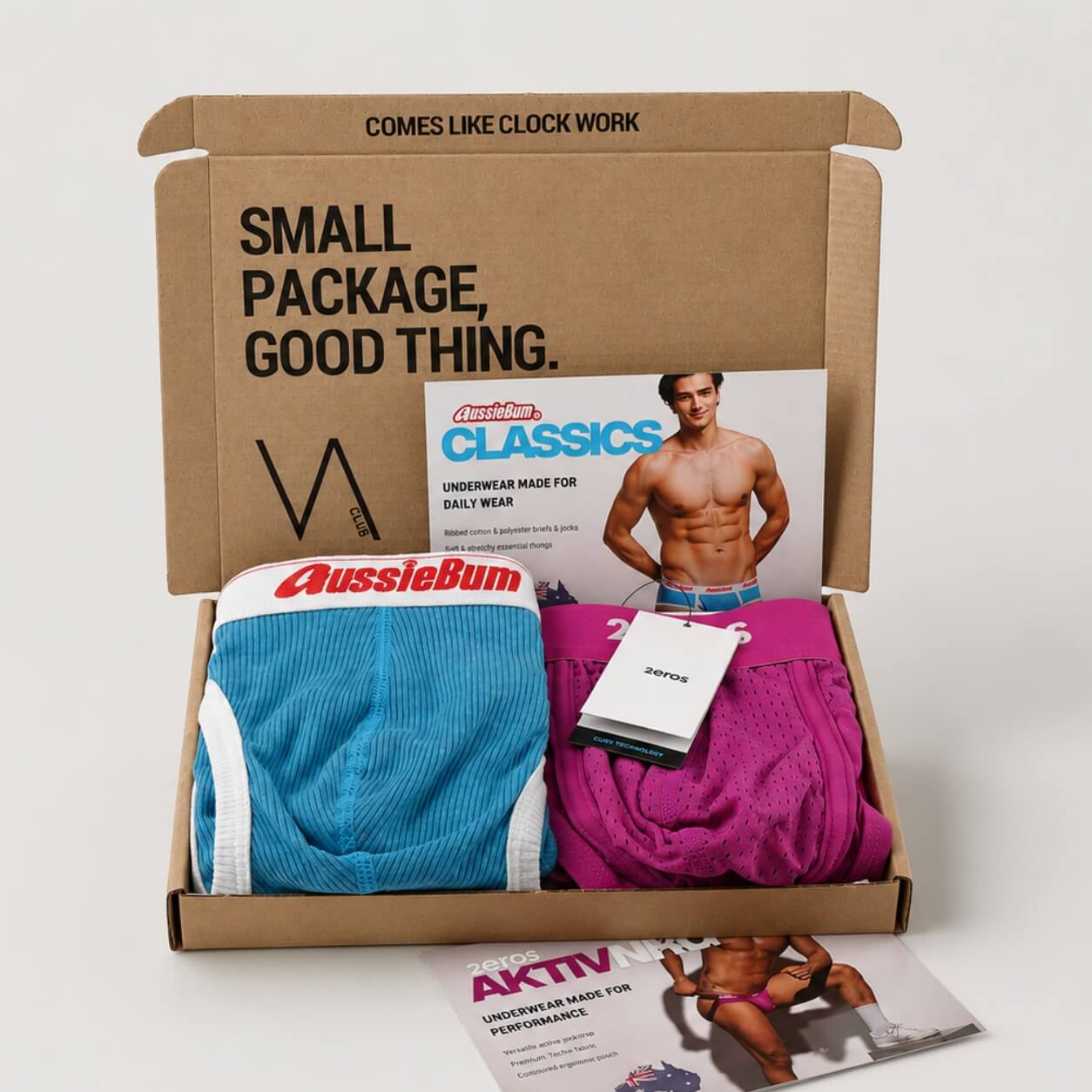 An example of a VA CLUB subscription cardboard box on a light grey background containing a 2eros fuchsia pink mesh brief and an aussieBum classic blue ribbed cotton brief, with brand tags, VOCLA branded tissue paper and a small printed product insert card