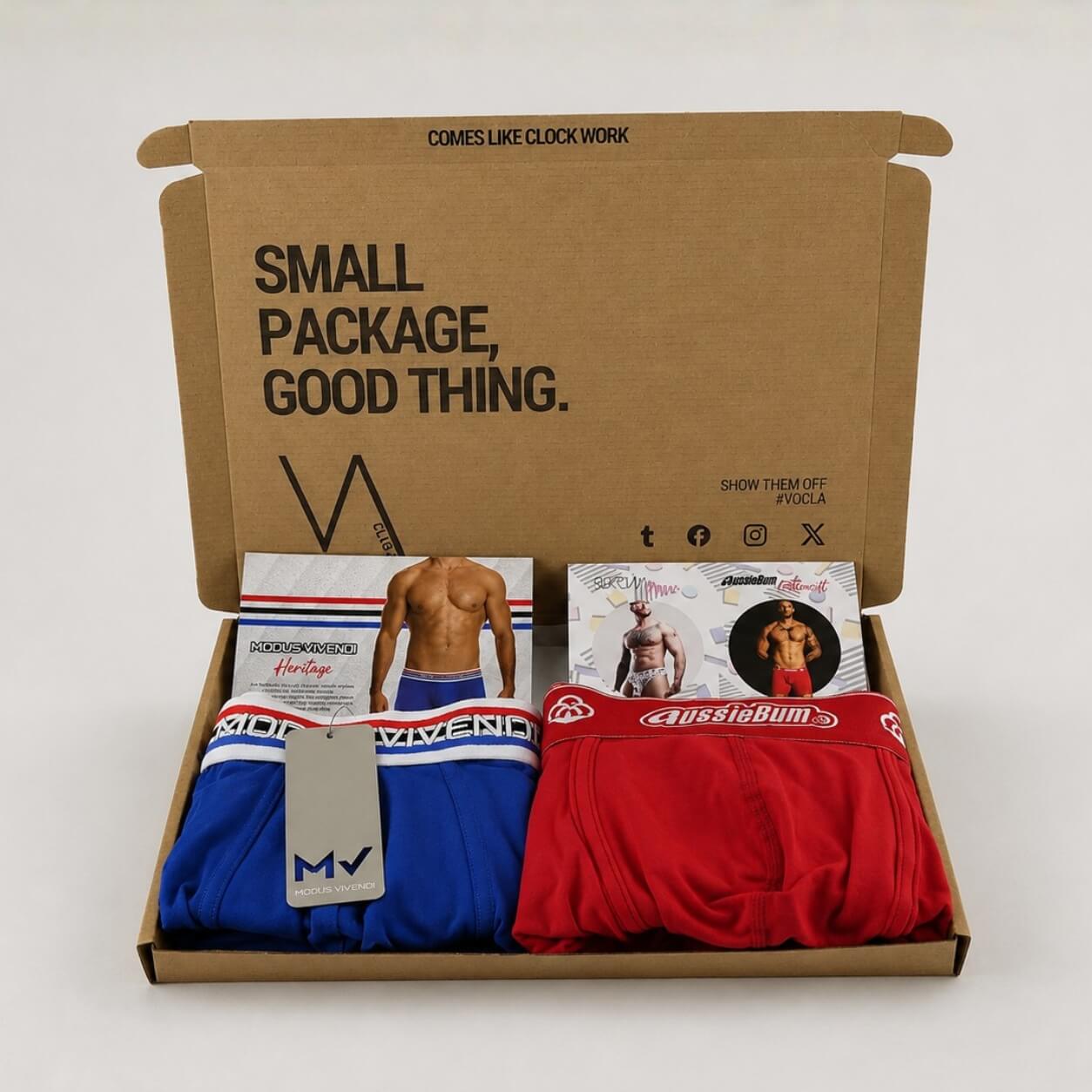 An example of a VA CLUB men's subscription box on a light grey background containing a MODUS VIVENDI royal blue Heritage boxer brief and an aussieBum red cottonsoft boxer trunk, with brand tags, VOCLA branded tissue paper and a small printed product insert card