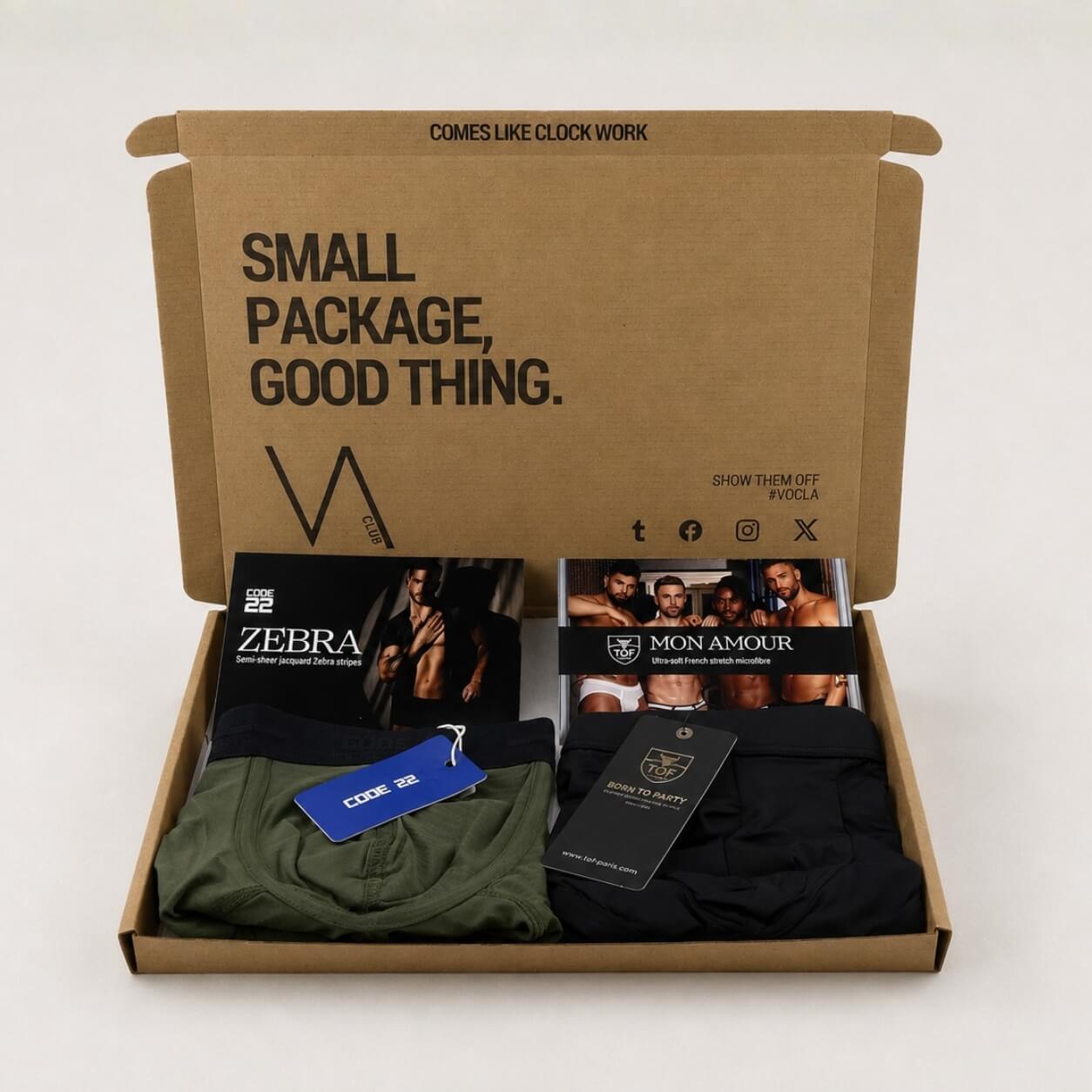 An example of a men's VA CLUB subscription box on a light grey background containing a CODE22 khaki Zebra brief and a TOF Paris Mon Amour black modal boxer trunk, with brand tags, VOCLA branded tissue paper and a small printed product insert card