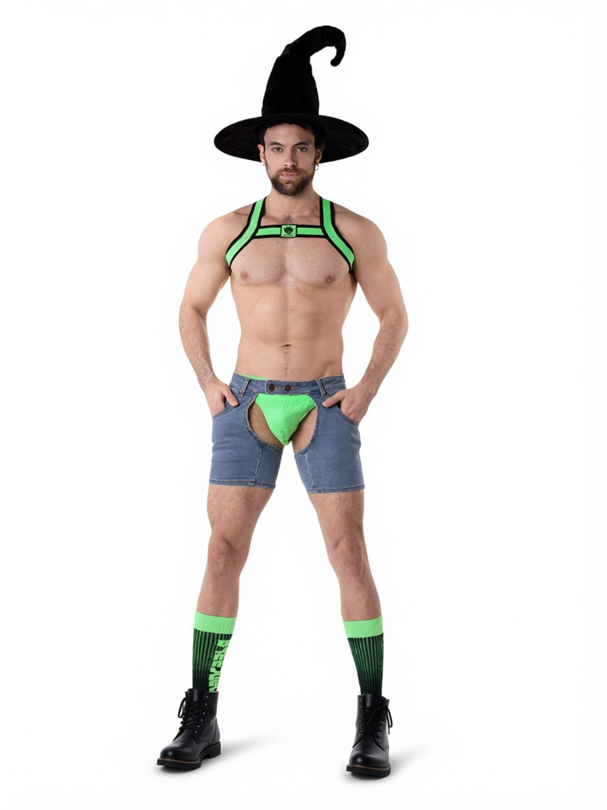 A man modeling a green and black witch-themed harness vest, denim shorts, green and black socks, and a black witch hat against a plain white background.