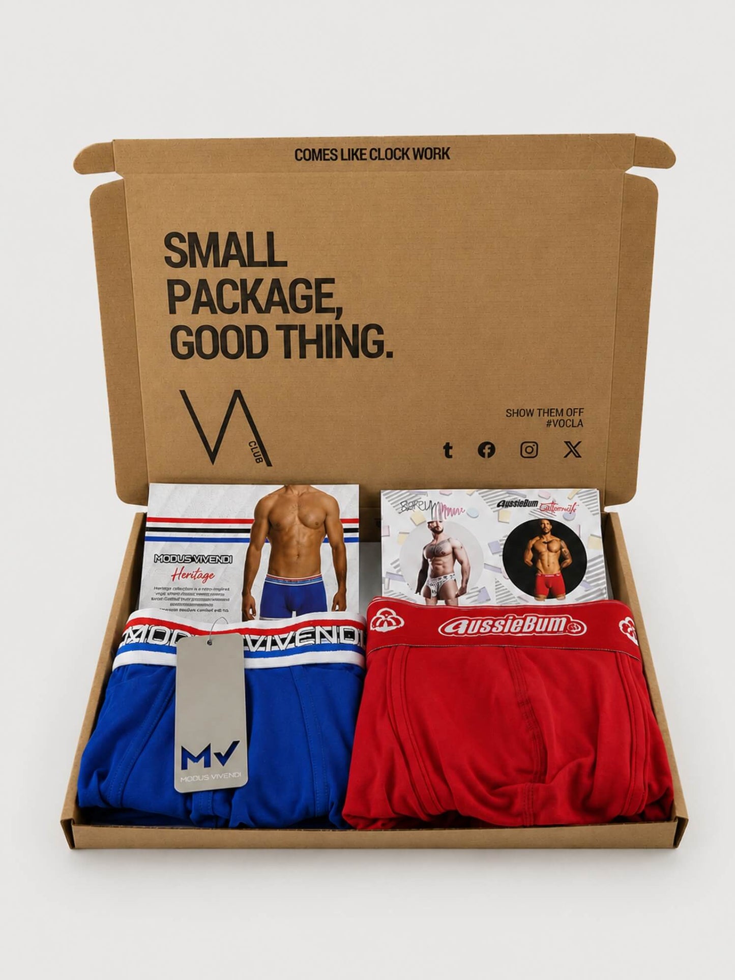 An open brown cardboard VA CLUB subscription box on a light grey background, containing folded men's underwear in blue and red, plus printed inserts and a small model photo. The box lid has printed text including 'SMALL PACKAGE, GOOD THING.