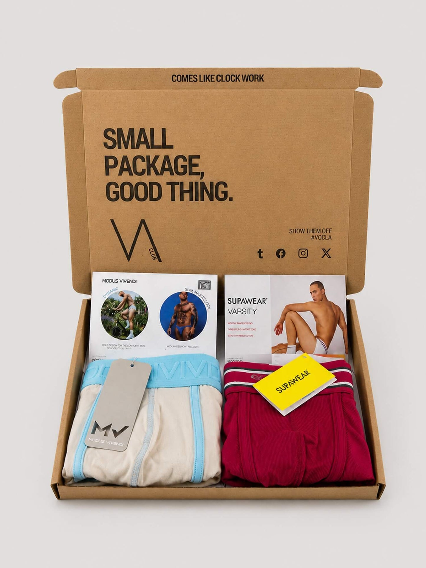 An open brown cardboard VA CLUB subscription box on a light grey background, containing folded underwear in cream and light blue, and burgundy, with printed inserts and a yellow tag. The inside lid reads 'SMALL PACKAGE, GOOD THING.