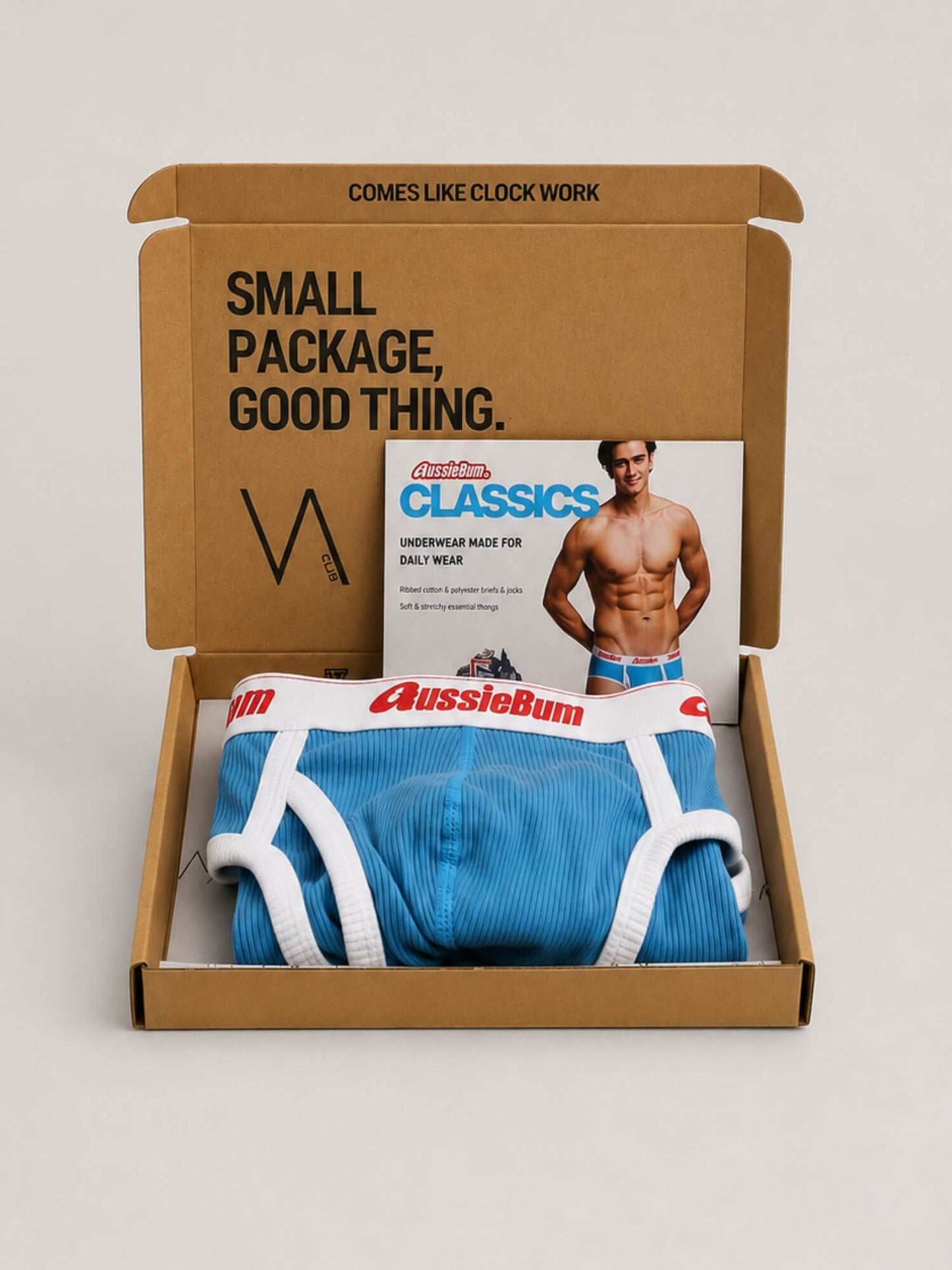 A brown cardboard gift box opened on a light grey background, containing folded blue and white Aussiebum brief underwear and a printed insert card featuring a shirtless male model. The box lid has printed text reading 'SMALL PACKAGE, GOOD THING.