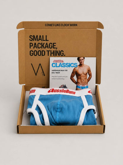 A brown cardboard gift box opened on a light grey background, containing folded blue and white Aussiebum brief underwear and a printed insert card featuring a shirtless male model. The box lid has printed text reading 'SMALL PACKAGE, GOOD THING.