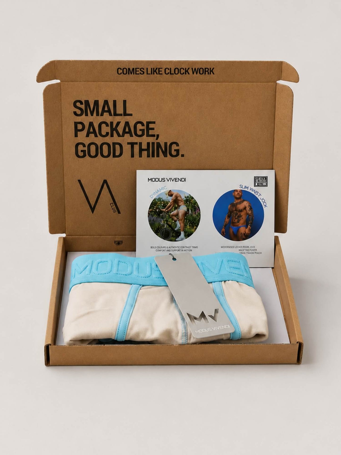 An open brown cardboard subscription box on a light grey background containing folded underwear in light blue and off-white, a branded card, and a hangtag. The box lid is printed with the text 'SMALL PACKAGE, GOOD THING.
