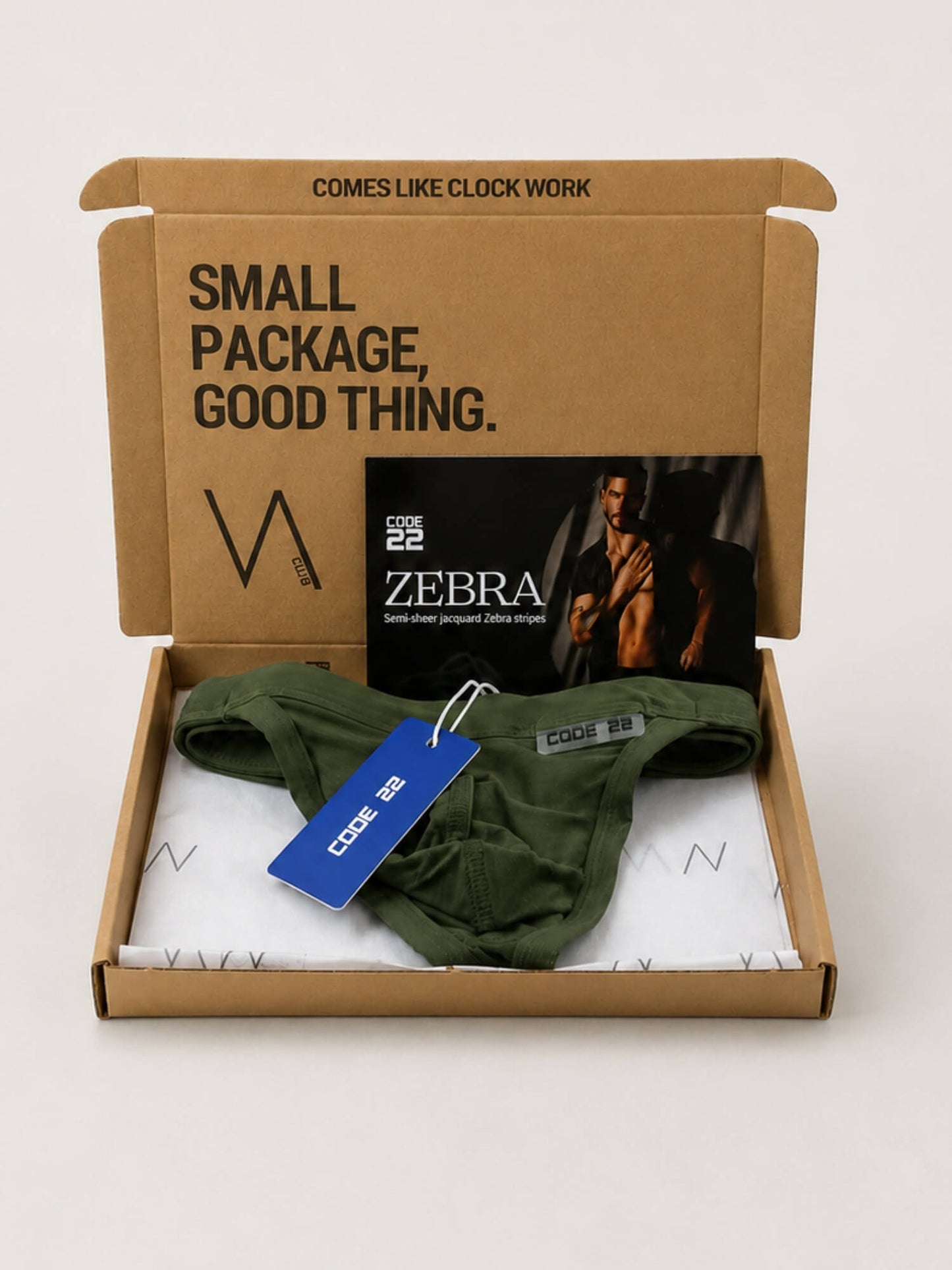 An open cardboard VA CLUB subscription box on a light neutral background containing an olive green pair of underwear thongs, a blue CODE 22 hangtag, and a ZEBRA-branded card. The box lid has printed text reading 'SMALL PACKAGE, GOOD THING.