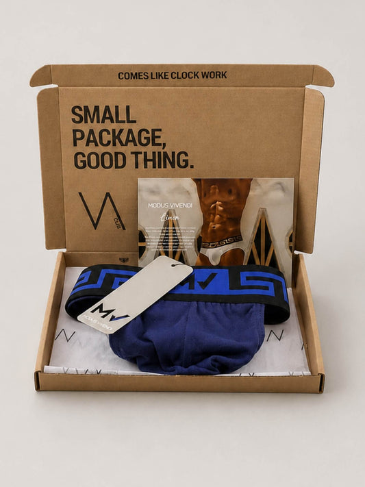 A brown cardboard VA CLUB subscription box opened on a light grey background, containing a MODUS VIVENDI navy blue jockstrap with a blue and black Greek key waistband, a white hangtag, and a small booklet. The inside lid has black printed text including 'SMALL PACKAGE, GOOD THING.