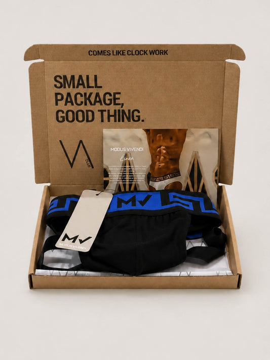 An open kraft cardboard VA CLUB subscription box on a light grey background, containing a black MODUS VIVENDI jockstrap and a beige hangtag. The box lid is printed with the text 'SMALL PACKAGE, GOOD THING.