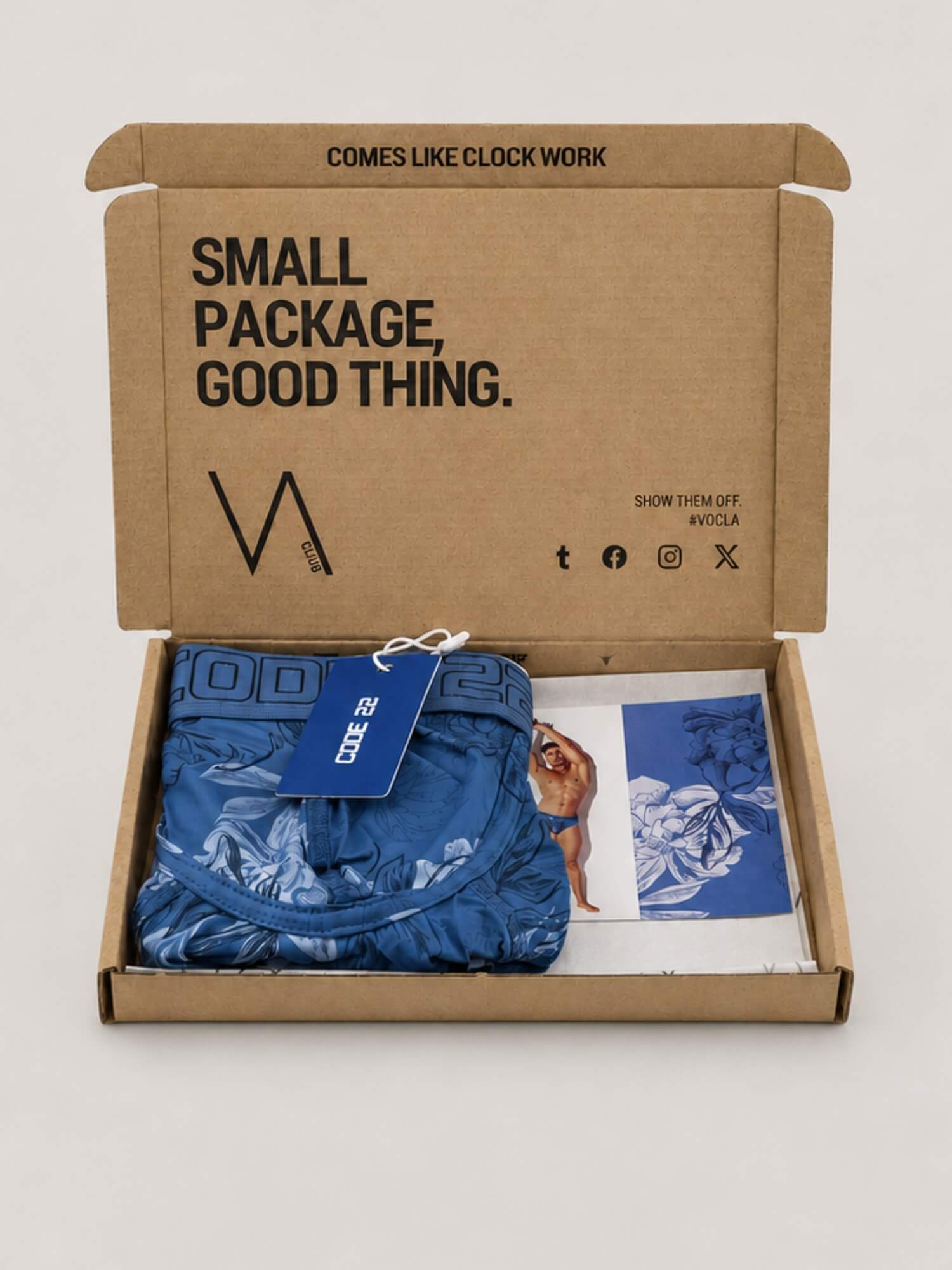 An open kraft cardboard subscription box on a light grey background, containing folded blue floral patterned underwear with a blue CODE 22 brand tag, a printed card, and branded interior text reading 'SMALL PACKAGE, GOOD THING.'.
