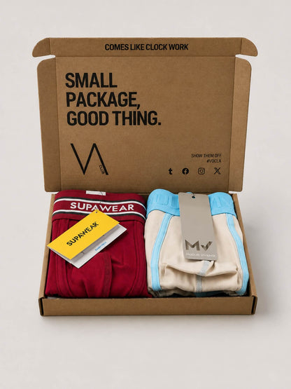 An open brown cardboard VA CLUB subscription box on a light grey background containing folded underwear. The left folded pair is dark red with a white band reading SUPAWEAR and a yellow hangtag, and the right folded pair is cream with light blue trim and a gray hangtag.