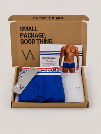 A brown cardboard gift box opened on a light grey background, containing folded blue MODUS VIVENDI boxer briefs with a white and blue striped waistband, a branded tag, a printed insert card showing a male model, and branded tissue paper. The box lid has printed text reading 'SMALL PACKAGE, GOOD THING.