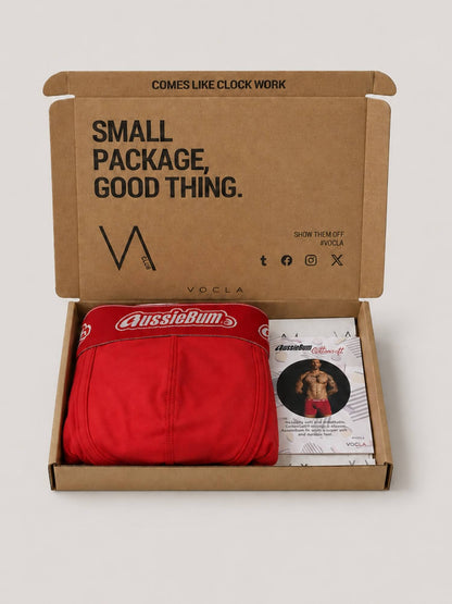 An open brown cardboard gift box on a light grey background, containing a folded red pair of AussieRum boxer briefs and a printed booklet. The inside lid reads 'SMALL PACKAGE, GOOD THING.
