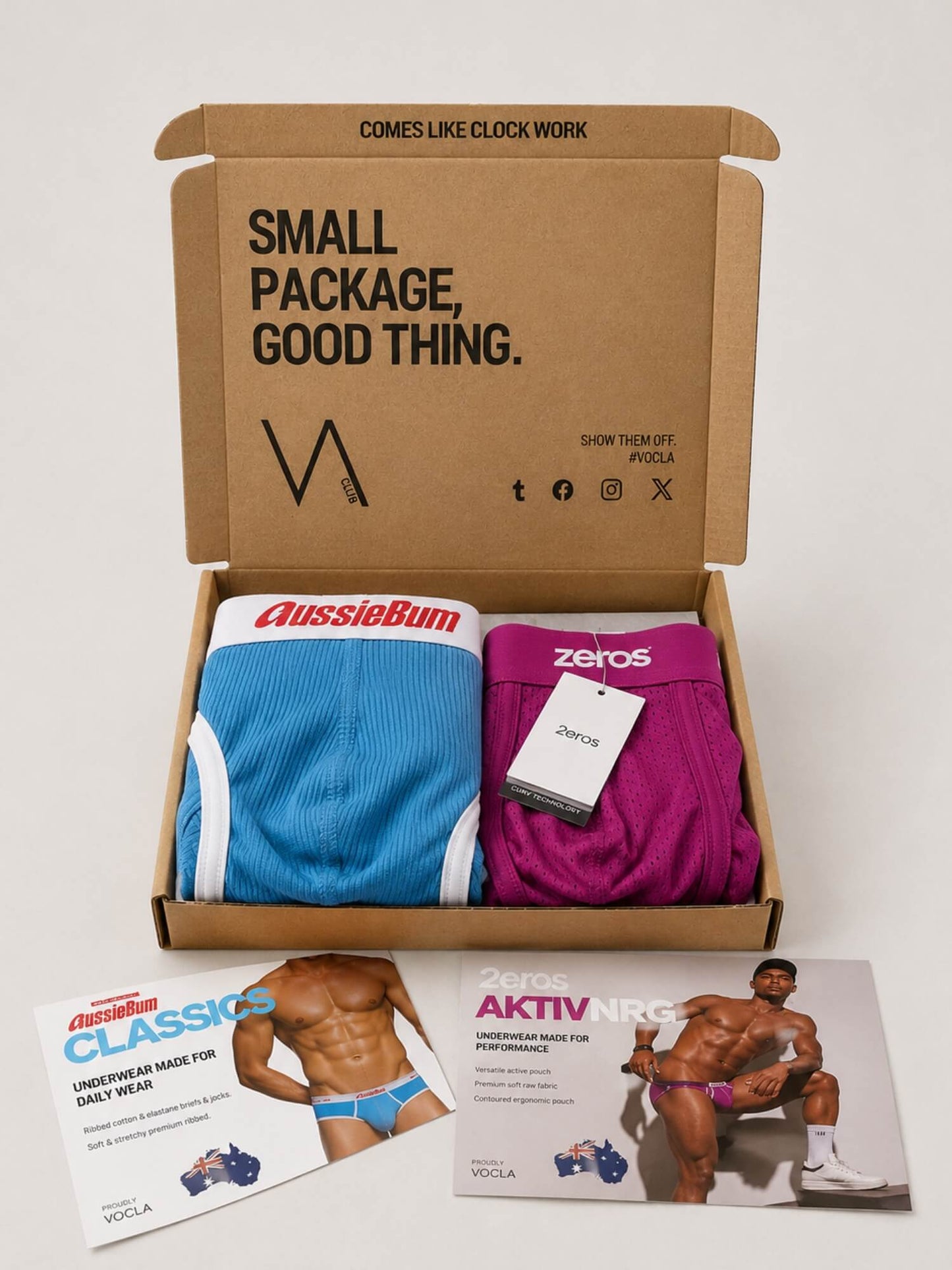 An open cardboard VA CLUB subscription box containing two pairs of underwear: a blue pair with white trim labelled 'aussieBum' and a fuchsia pink pair labelled '2eros'. Two printed inserts with photos and text are placed in front on a light grey background.