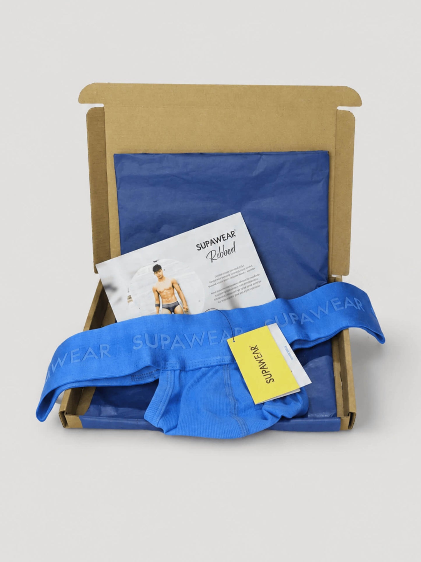 A blue pair of men's thongs from SUPAWEAR is displayed inside an open brown cardboard box with a blue insert, alongside a branded paper insert and a yellow hangtag.