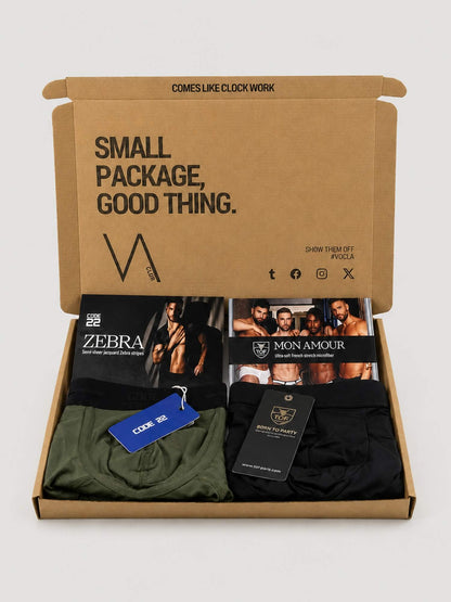 An open brown cardboard subscription box on a light grey background, containing folded men's underwear and printed inserts. Visible products include olive green and black CODE 22 briefs with blue and black hang tag, and TOF PARIS MON AMOUR underwear. 