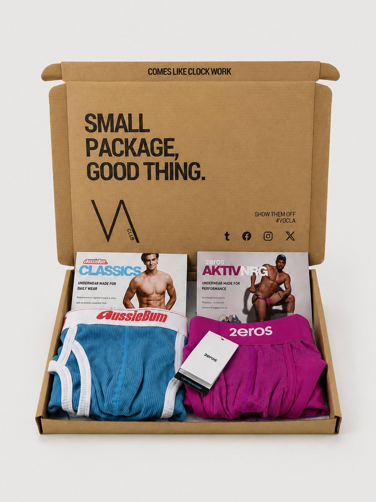 An open brown VA CLUB cardboard subscription box containing folded underwear and printed inserts. Visible products include a blue pair with white trim labelled AussieBum, a magenta pair labelled 2eros, and two magazine-style inserts, all arranged inside a branded box on a light grey background.