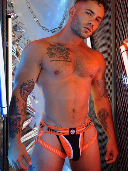 Man wearing a black and orange jockstrap with tattoos visible on his arms and chest.