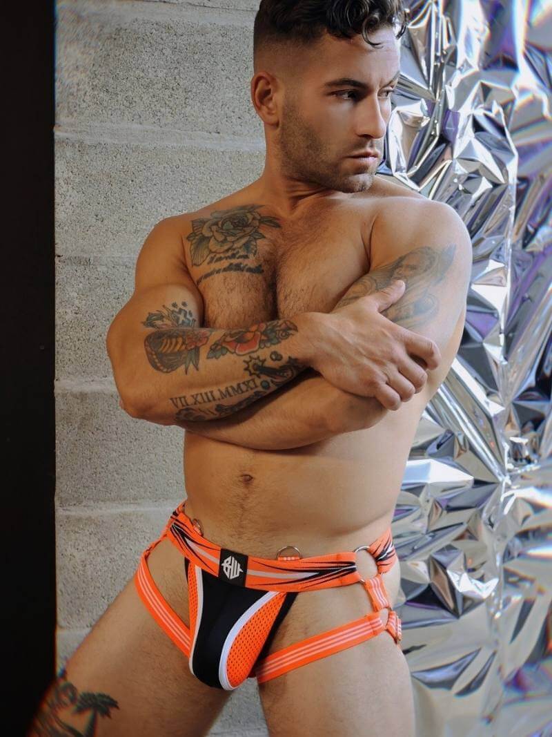 Man wearing a colorful clubwear jockstrap with tattoos on his arms and chest, standing against a reflective surface.
