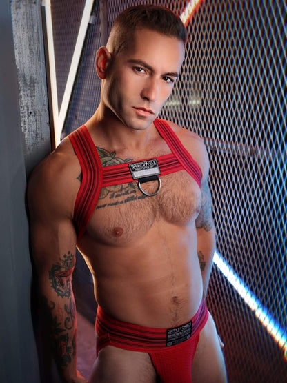 Man wearing red harness and underwear against a textured wall.